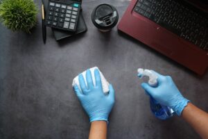 A person cleaning a computer keyboard and screen with a microfiber cloth, surrounded by cleaning supplies.